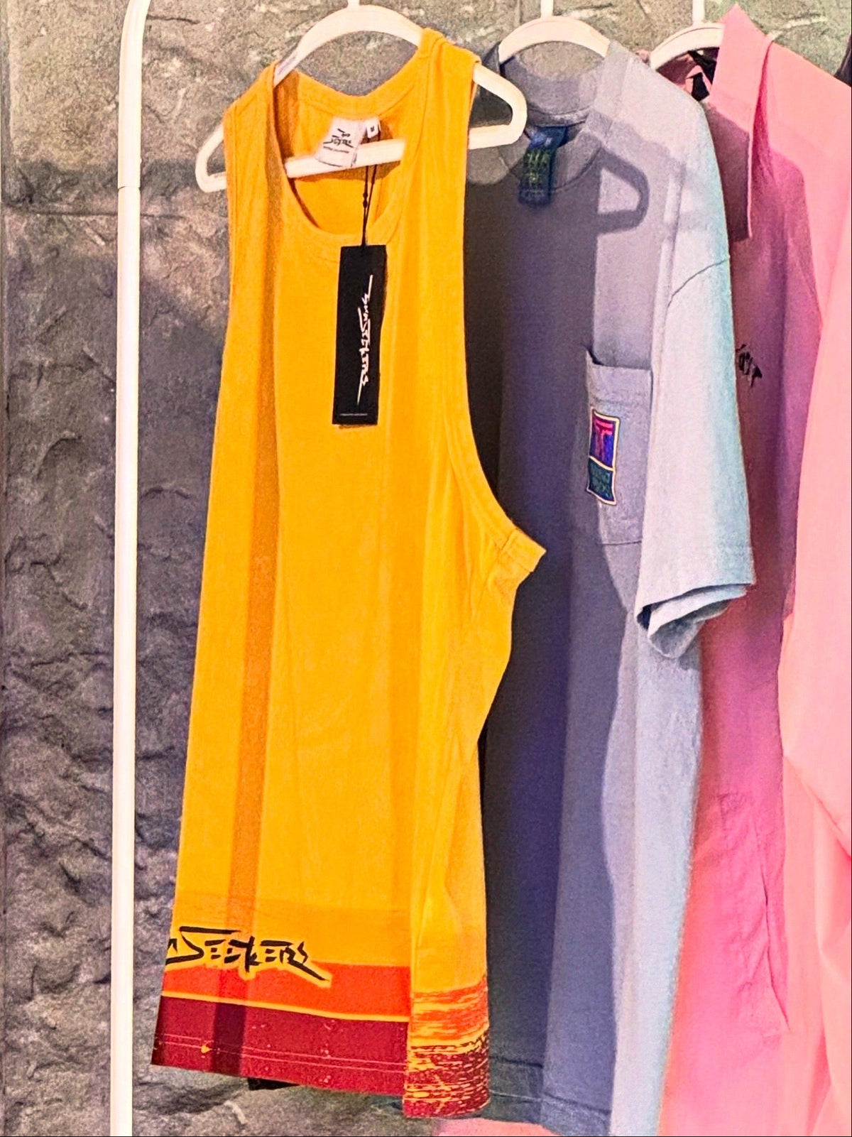 A bright yellow sleeveless tank top with a red and orange graphic hem, hanging on a white hanger next to a light blue pocket tee and a pink top against a textured gray wall.