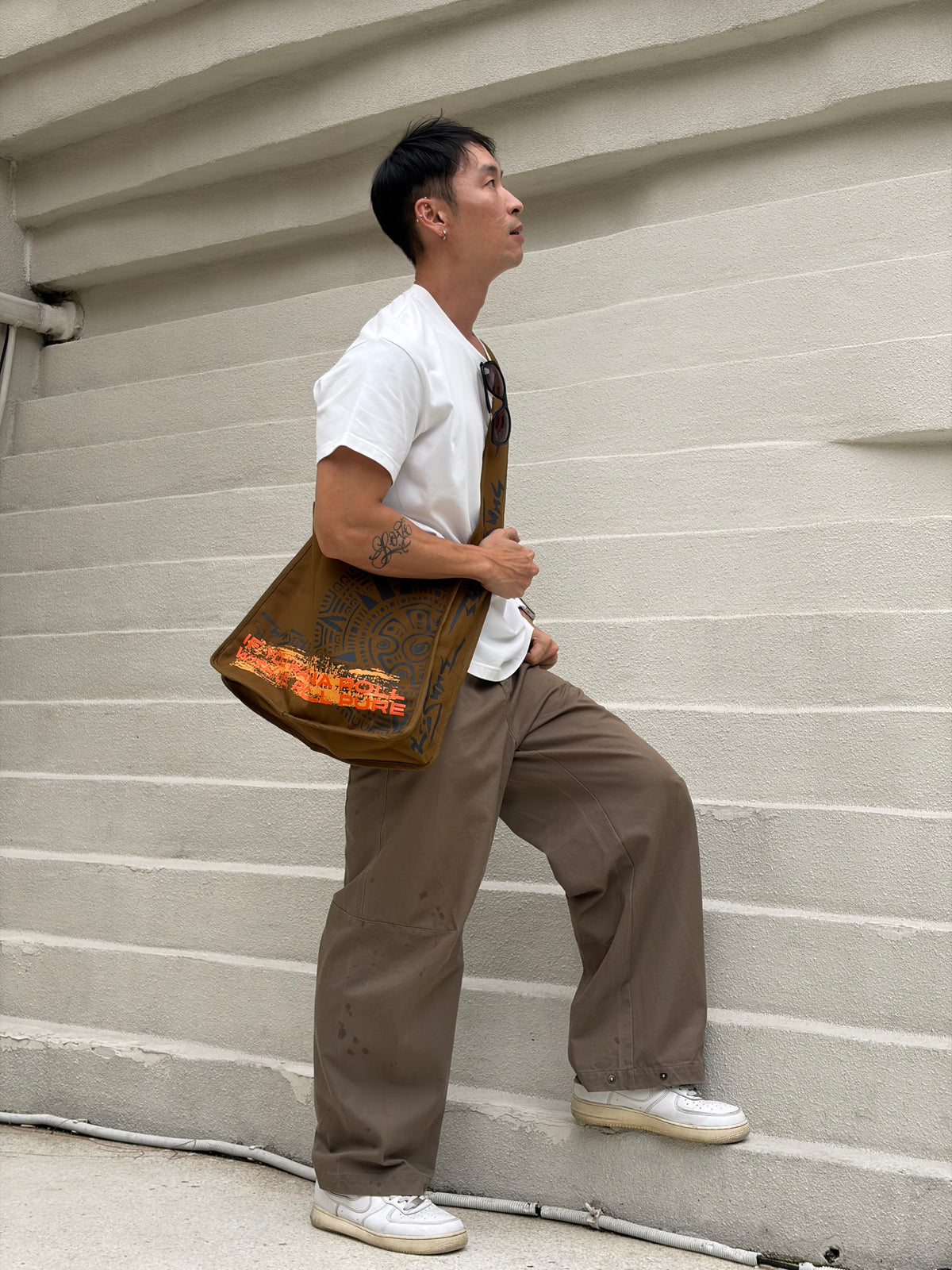A man wearing Sunseekers Khaki messenger bag worn crossbody and a white t-shirt.